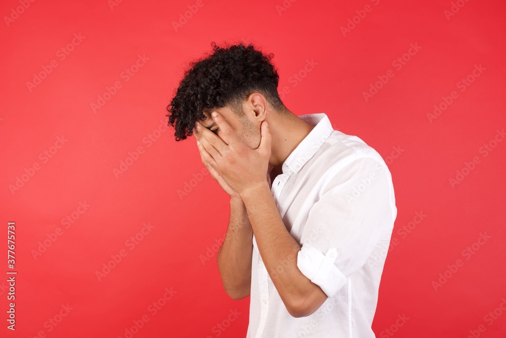 Sad Young arab man with afro hair wearing shirt standing over isolated red background crying covering his face with his hands.