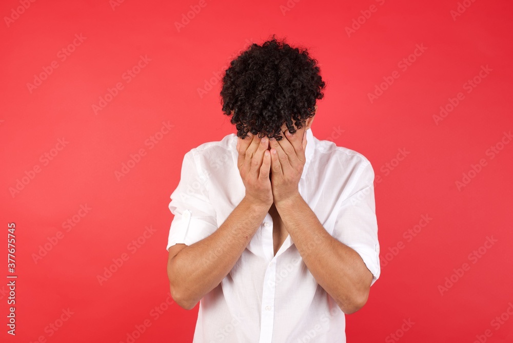 Fototapeta premium Young arab man with afro hair wearing shirt standing over isolated red background covering her face with his hands, being devastated and crying.