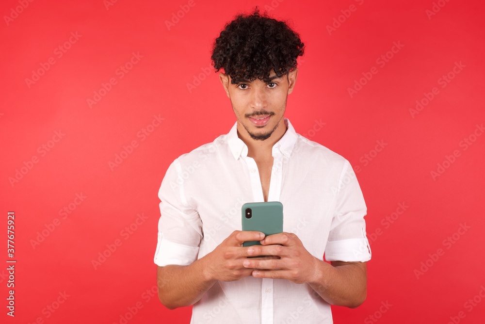 Young arab man with afro hair wearing shirt standing over isolated red wall enjoys distant communication, uses mobile phone, surfs fast unlimited internet, has pleasant smile,  makes shopping online, 
