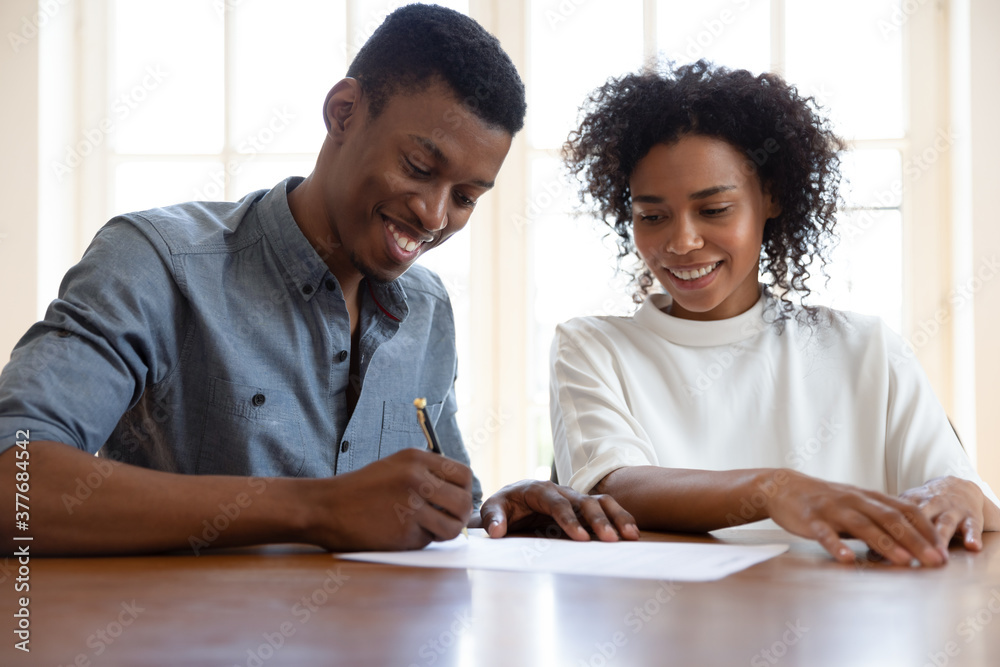 Obraz premium Excited African American young couple sit at desk put signature on contract close deal with realtor or broker, happy biracial husband and wife sign document agreement buying first house together