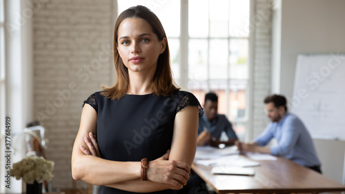 Headshot portrait of focused Caucasian young businesswoman stand forefront posing at company office meeting, serious millennial female boss or CEO look at camera show confidence and success