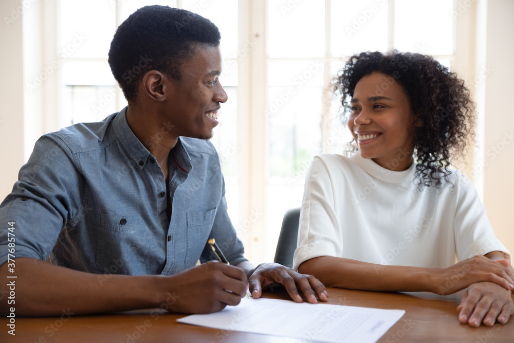 Excited young couple sit at desk put signature on contract feel euphoric buying first house together, smiling African American husband and wife look at each other sign agreement at realtor office Excited young couple sit at desk put signature on contract feel euphoric buying first house together, smiling African American husband and wife look at each other sign agreement at realtor office