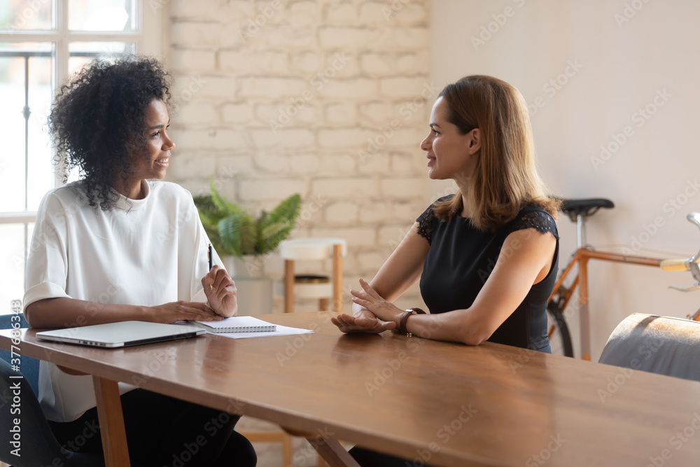 Smiling African American young businesswoman talk negotiate with female Caucasian job candidate at office meeting, happy biracial boss discuss ideas with business partner or employee at interview Smiling African American young businesswoman talk negotiate with female Caucasian job candidate at office meeting, happy biracial boss discuss ideas with business partner or employee at interview