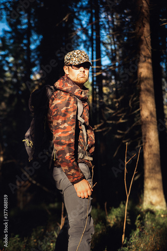 Hiker hiking in autumn forest. Male hiker in camouflage jacket with backpack looking to the side walking in forest. Caucasian handsome male outdoors in nature. Concept of forest wear, navigation.
