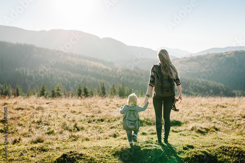 Mom with backpack and child walk in autumn grass. The daughter and mother walking on nature. Family spending time together in mountain on vacation. Holiday trip concept. World Tourism Day. Back view.