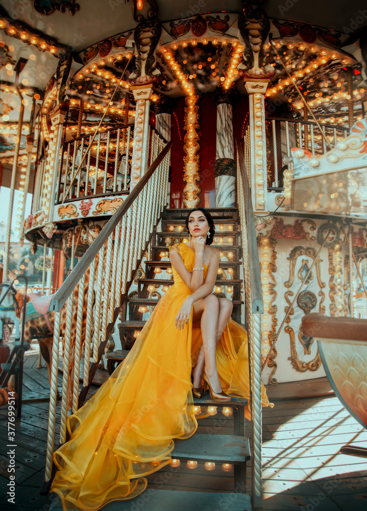 Young beautiful woman in a bright yellow evening dress sitting on stairs. Fashion model posing against the backdrop of an amusement park and carousel