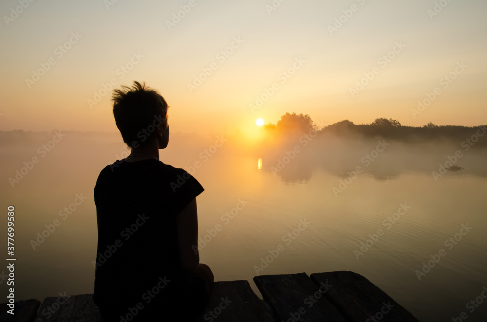 Silhouette of a girl sitting on the pier against the sunset sky with copy space. The girl looking at the rising sun over the lake in the fog. Reflection of the sun in the water surface.