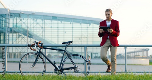 Caucasian young stylish handsome man standing at street with bike, videochatting on tablet device. Male having talk via webcam online on gadget computer with bicycle outside. Videochatting talking.