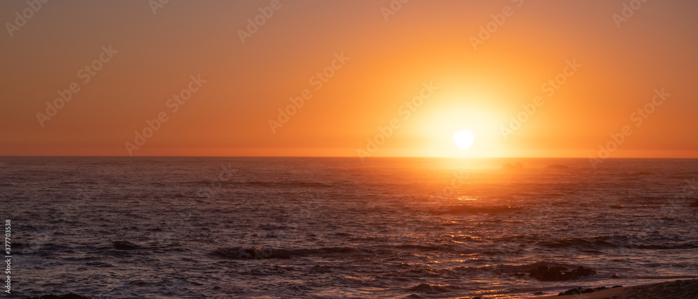 Bright vivid sun on horizon over ocean at sunset, with reflections on water. Wide aspect ration with copy space on left.