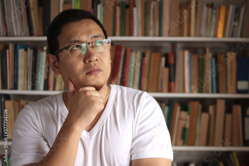 Asian man thinking while standing in front of bookshelf, education concept, studying in library, serious expression