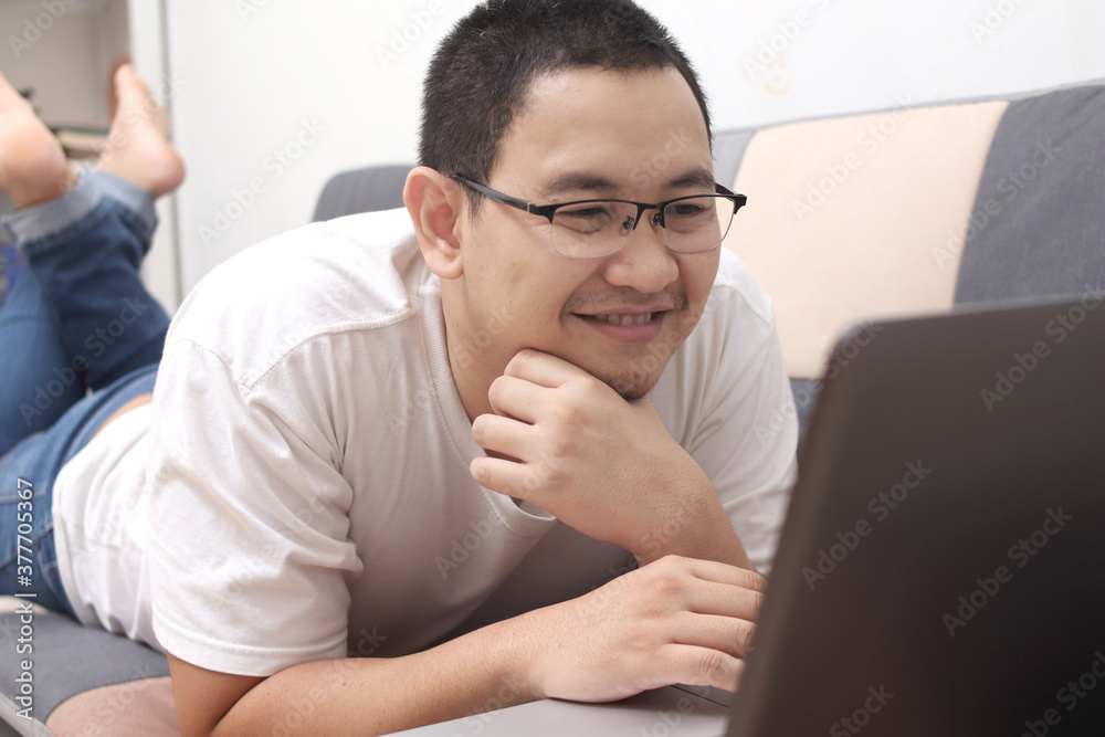 Young Asian man smiling while looking at his laptop on sofa Young Asian man smiling while looking at his laptop on sofa