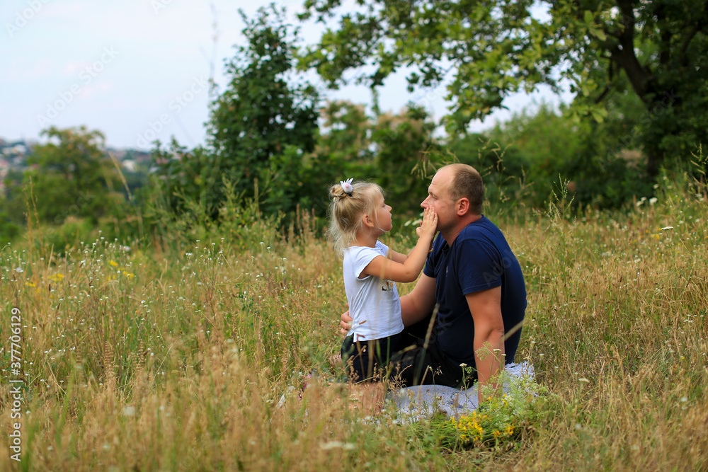 A father with a three-year-old blonde daughter communicate warmly while sitting on the grass in nature in the field grasses. The daughter whispers something in her ear gently. Father's day. 