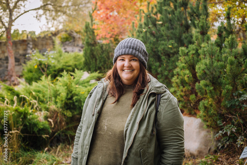 Young  cheerful curvy woman in grey hat and green jacket walking in the park