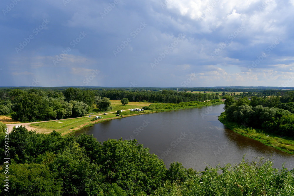 A view from the top of a hill with a vast yet shallow river or lake flowing through some forests and moors with a cloudy sky right before the thunderstorm visible in the distance seen in Poland