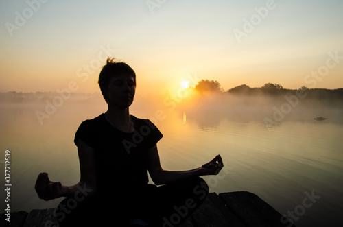 Silhouette of a meditating girl against the background of sunrise with copy space. Woman in kubera mudra meditation. Reflection of the sun in the water surface.