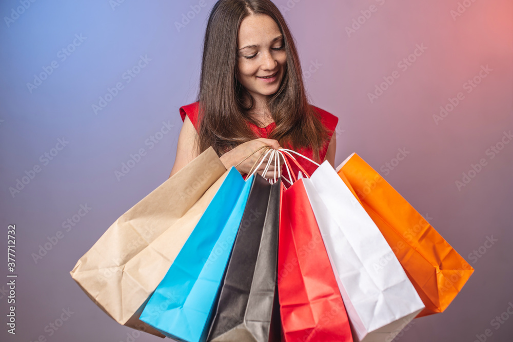 Young woman in a red dress is holding a lot of multicolored paper bags with purchases. Concept of shopping and sales. Blue and pink background