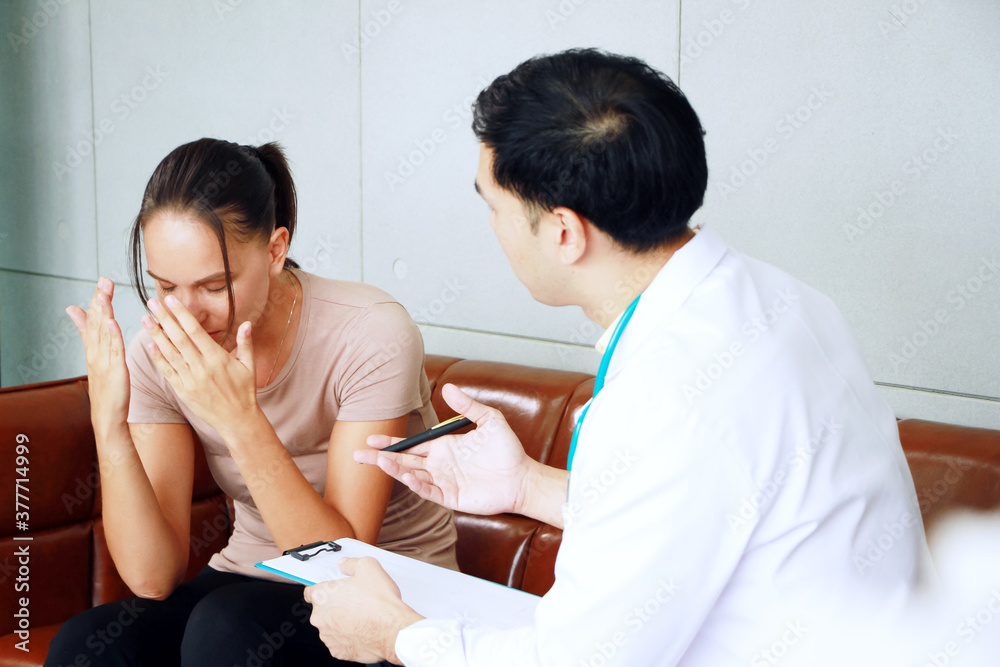 Young asian male psychiatrist is specialist in psychiatry is talking with Caucasian female Patient is headache and encouraging to her in Clinical treatment for emotional symptoms. focus at a woman. Young asian male psychiatrist is specialist in psychiatry is talking with Caucasian female Patient is headache and encouraging to her in Clinical treatment for emotional symptoms. focus at a woman.