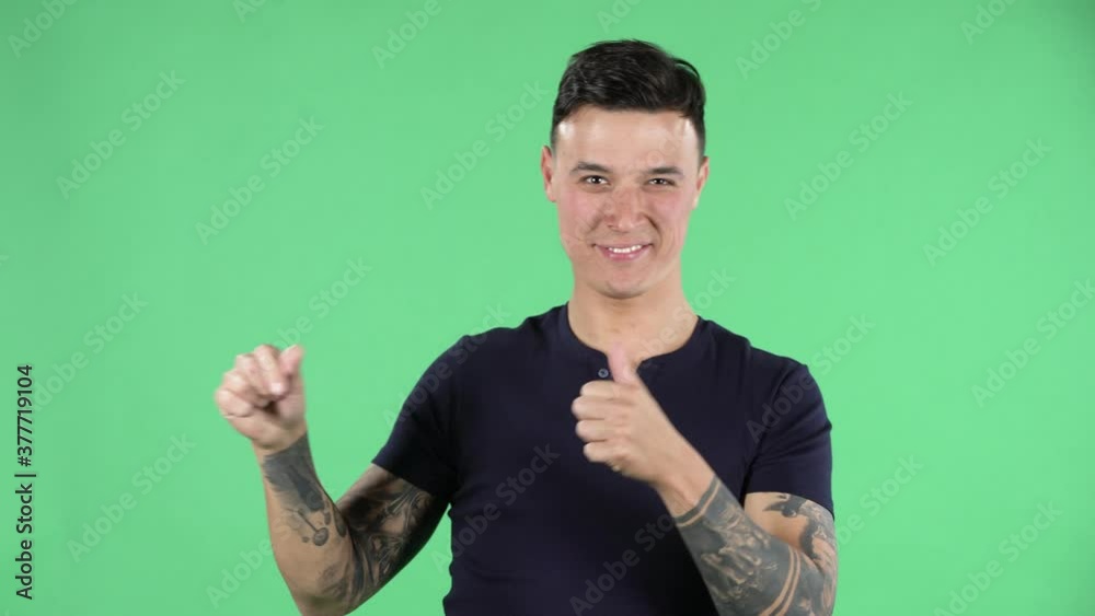 Portrait of a handsome young guy is looking at camera and pointing side hand for something then shows thumb like. Green screen.