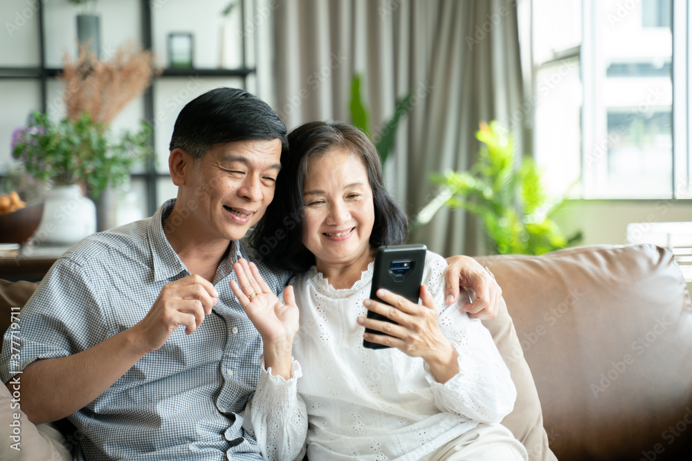 Social distancing or new normal concept.Senior asian couple sitting on sofa in home playing smartphone and video conference with grandchild.They are smiling to spend their time together with happiness Social distancing or new normal concept.Senior asian couple sitting on sofa in home playing smartphone and video conference with grandchild.They are smiling to spend their time together with happiness