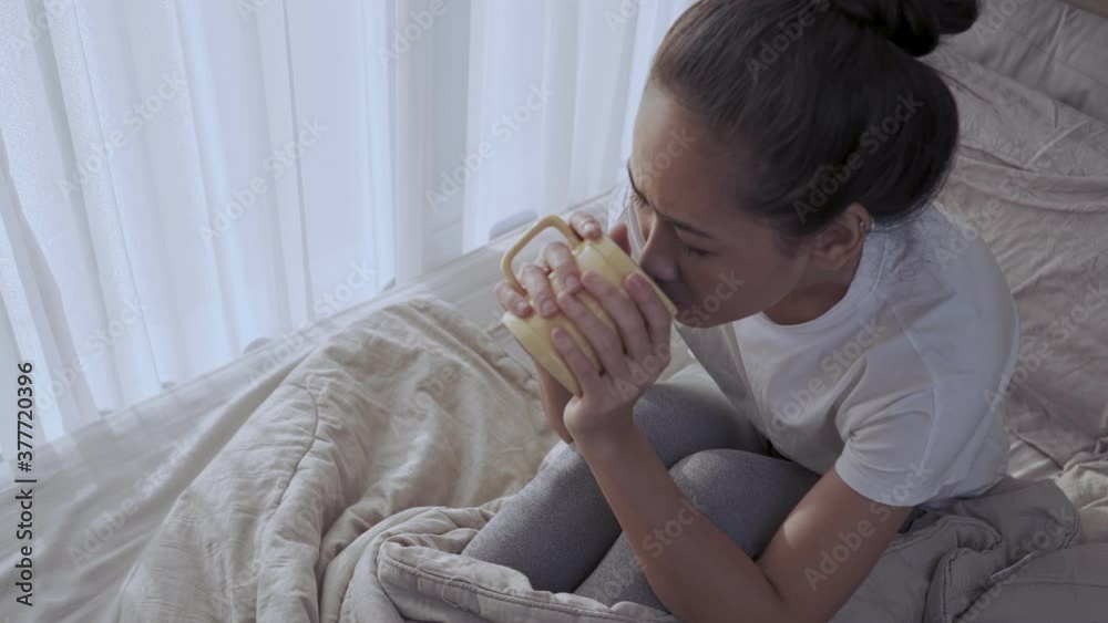Above shot tan skin Asian woman Drinking hot beverage from coffee mug, express sad and loneliness emotion, sit down alone on the bed, inside comfortable messy bedroom, isolated woman covid pandemic Above shot tan skin Asian woman Drinking hot beverage from coffee mug, express sad and loneliness emotion, sit down alone on the bed, inside comfortable messy bedroom, isolated woman covid pandemic
