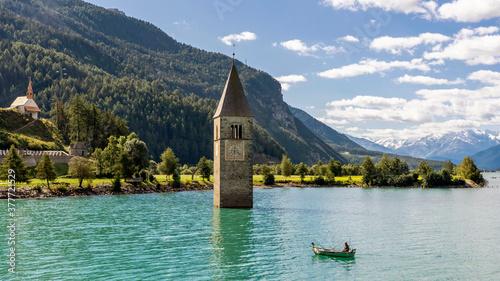 Fototapeta Naklejka Na Ścianę i Meble -  Fisherman in his boat near the submerged bell tower in Resia lake, Curon, South Tyrol, Italy