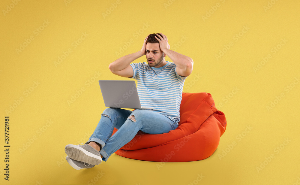 Worried young man with laptop on bean bag chair, yellow background Worried young man with laptop on bean bag chair, yellow background