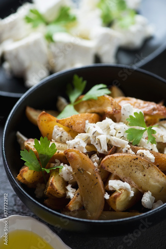 Closeup of a black bowl with baked potato wedges served with feta cheese in a greek style, vertical shot, selective focus