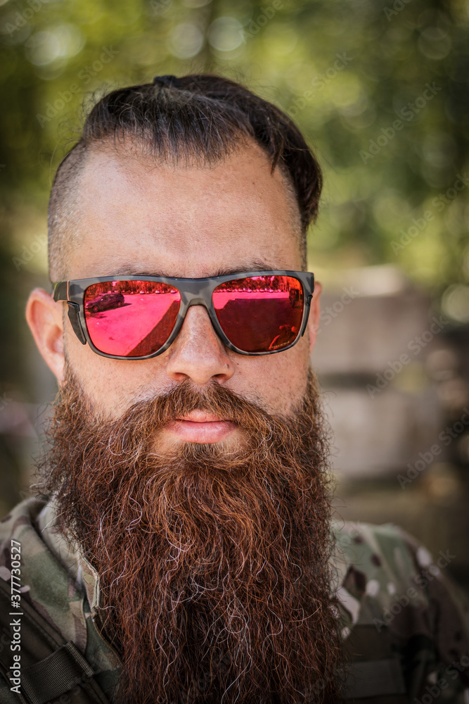 Bearded man with sunglasses in the military desert uniform and body armor stands and reloads his cartridge. The bearded player in the airsoft reloads his carabine. Outside airsoft. Tattooed arms Bearded man with sunglasses in the military desert uniform and body armor stands and reloads his cartridge. The bearded player in the airsoft reloads his carabine. Outside airsoft. Tattooed arms