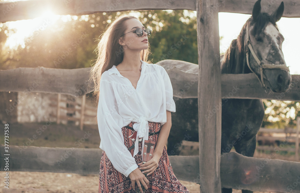 a woman of model appearance with long hair is dressed in a shirt with a long skirt and holds a guitar in her hands stands near the fence against the background of an equestrian farm near the horses
a woman of model appearance with long hair is dressed in a shirt with a long skirt and holds a guitar in her hands stands near the fence against the background of an equestrian farm near the horses