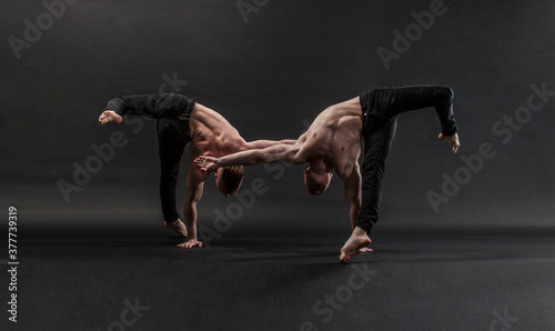 two twin brothers in black jeans with a naked torso perform acrobatic elements, black background 