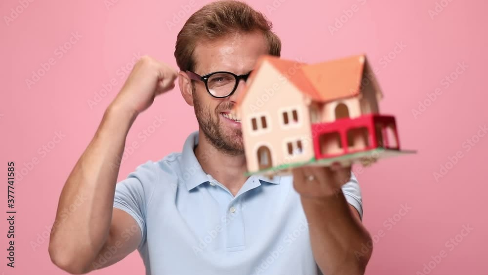 casual young man in blue polo shirt recommending house model, smiling, holding fists in the air and celebrating, laughing and screaming on pink background casual young man in blue polo shirt recommending house model, smiling, holding fists in the air and celebrating, laughing and screaming on pink background