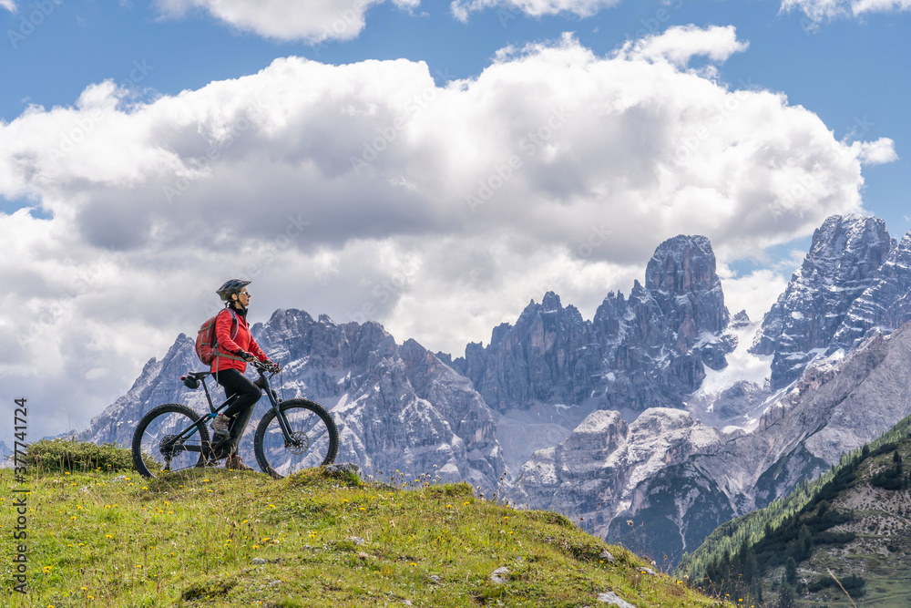 nice and active senior woman riding her electric mountain bike on the high plateau of Pratto Piazzo in the three peaks Dolomites , rocky silhouette of Mount Cristallo in background, South Tirol, Italy