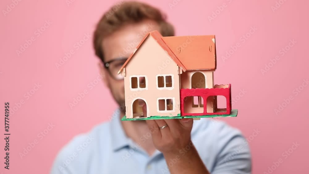 young casual man in blue polo shirt covering face with house model, showing and offering keys on pink background young casual man in blue polo shirt covering face with house model, showing and offering keys on pink background