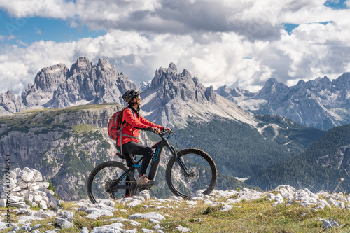 nice and active senior woman riding her electric mountain bike on the high plateau of Pratto Piazzo in the three peaks Dolomites , rocky silhouette of Mount Cristallo in background, South Tirol, Italy