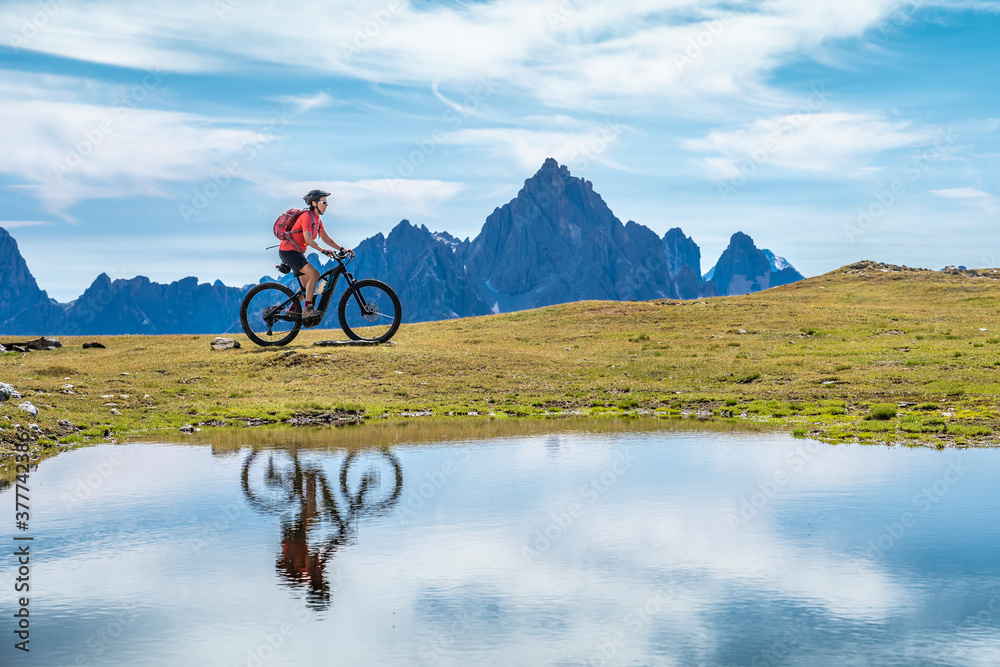 nice woman riding her electric mountain bike the Three Peaks Dolomites, reflecting herself in the blue water of a cold mountain lake