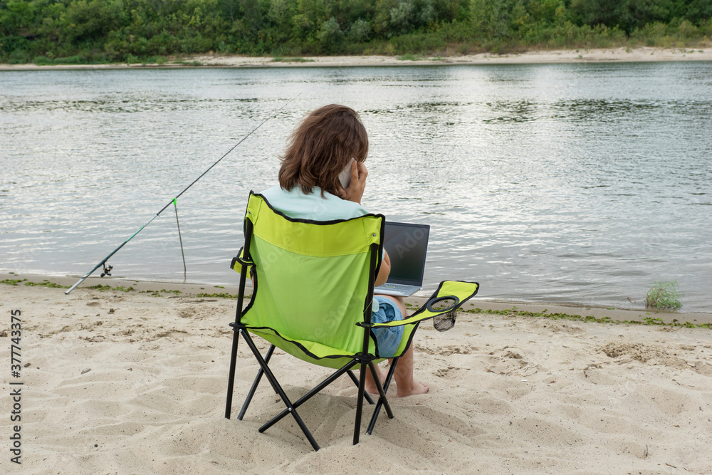 Back view of young female sit on bank of river with laptop on knees and smartphone in her hand. Copy space for text. Remote work or education concept. Back view of young female sit on bank of river with laptop on knees and smartphone in her hand. Copy space for text. Remote work or education concept.
