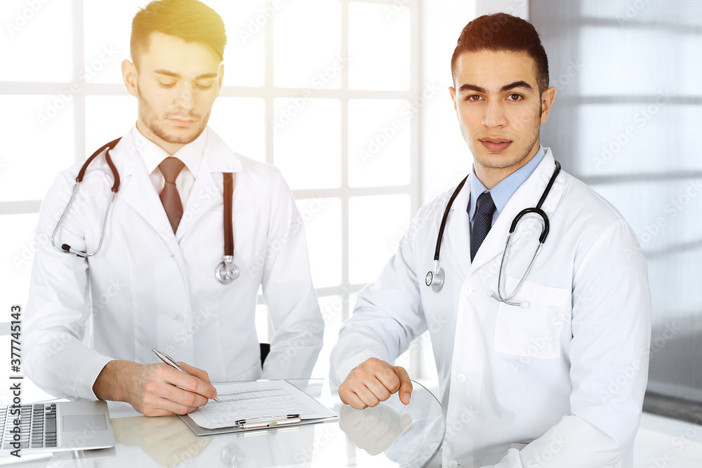 Fototapeta premium Arab doctor man sitting at the glass desk with caucasian colleague in medical office or sunny clinic. Diverse doctors team, medicine concept