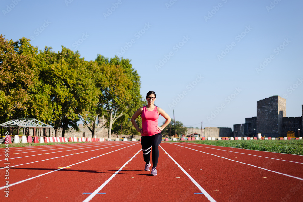 Active, 6 months pregnant woman workingout on track field on sunny morning. Attractive female doing walking lunges toward the camera.