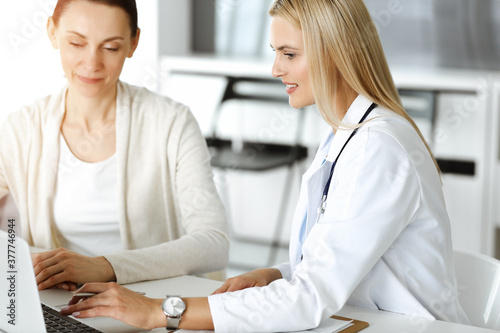 Woman-doctor at work in sunny hospital is happy to consult female patient. Blonde physician checks medical history record and exam results while using clipboard
