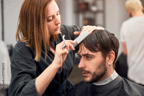 Handsome blue eyed man sitting in barber shop. Hairstylist Hairdresser Woman cutting his hair. Female barber.