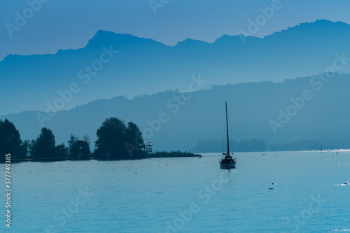 Sail boat along the shores of the Upper Zurich Lake (Obersee) with the Alps in the foggy morning background, Busskirch, Rapperswil-Joma, St. Gallen, Switzerland