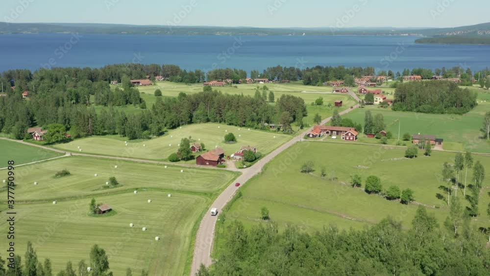 Motorhome caravan and single driving on small Swedish country road. Sweden countryside with red wooden cabins and summer fields and lake behind. Aerial drone video above forest trees