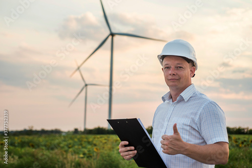 Windmill engineer in white helmet and with tablet shows thumb up on windmills background