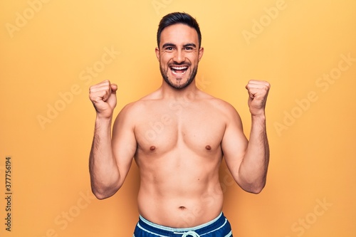 Young handsome man with beard wearing sleeveless t-shirt standing over yellow background screaming proud, celebrating victory and success very excited with raised arms