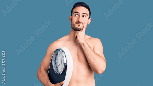 Young handsome man wearing swimwear holding weighing machine serious face thinking about question with hand on chin  thoughtful about confusing idea