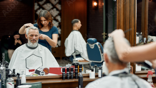 Handsome mature bearded man sitting in barbershop chair while female barber making haircut for him. Barbershop