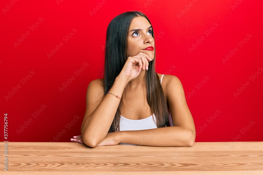 Young hispanic woman wearing casual clothes sitting on the table serious face thinking about question with hand on chin, thoughtful about confusing idea Young hispanic woman wearing casual clothes sitting on the table serious face thinking about question with hand on chin, thoughtful about confusing idea