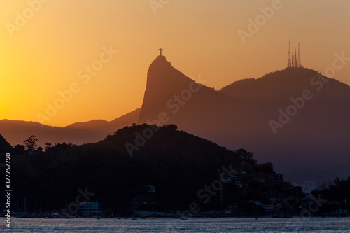 Beautiful distant view of Christ the Redeemer silhouette at sunset against orange sky; Mount Corcovado, Rio de Janeiro, Brazil. Smaller mountain and the sea in the foreground. Copy space for text.