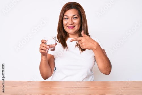 Middle age brunette hispanic woman drinking glass of water sitting on the table smiling happy pointing with hand and finger