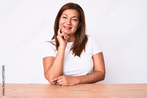 Middle age brunette hispanic woman wearing casual white tshirt sitting on the table smiling looking confident at the camera with crossed arms and hand on chin. thinking positive.